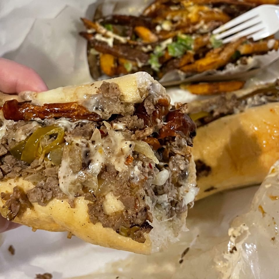 Hand holds a loaded cheesesteak sandwich with peppers and onions, alongside fries and another sandwich on foil.